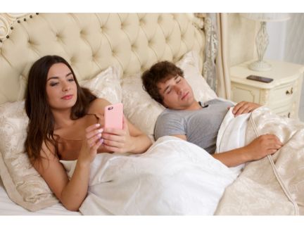 男性が寝る横でスマホを操作する女性の写真